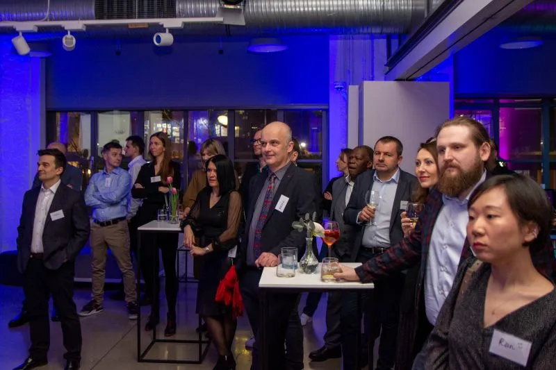MBA Event A corporate networking event in a modern venue with blue lighting. Several well-dressed professionals are standing in groups, holding drinks and engaged in conversation. Attendees are wearing name tags and gathered around small standing tables with drinks and small decorations. The atmosphere appears professional yet relaxed, with industrial-style ceiling elements visible above.