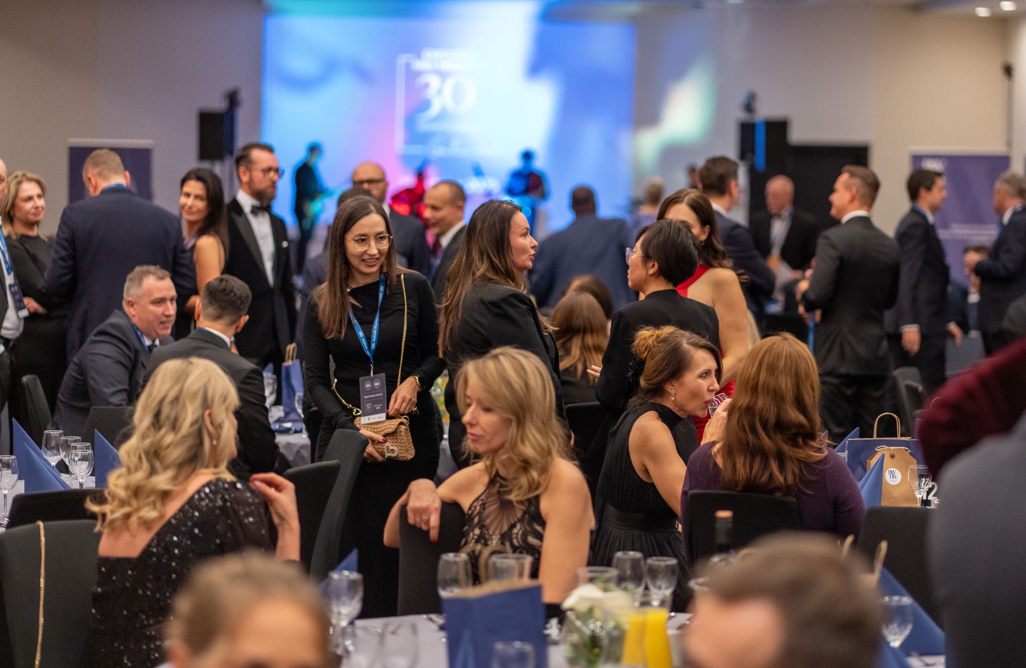Elegantly dressed guests chat at round tables during a formal gala or banquet; a stage with lighting and musical performance is visible in the background.