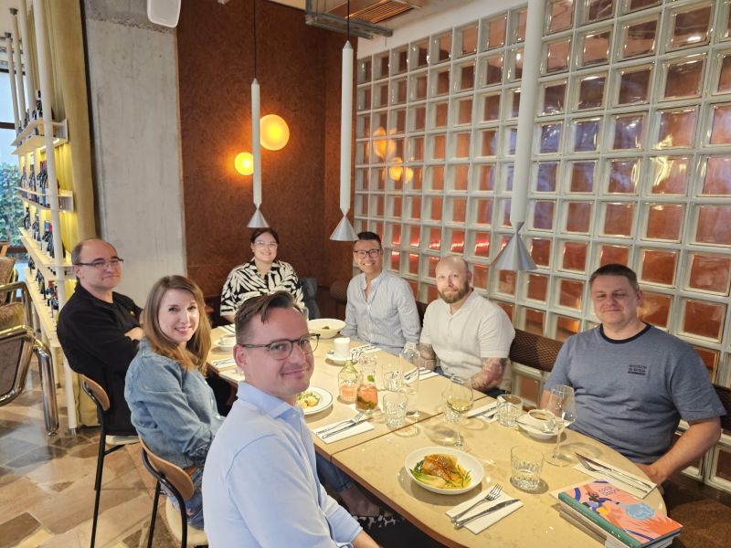 Seven people seated around a restaurant table during an informal book club meeting, with meals, drinks, and books on the table.